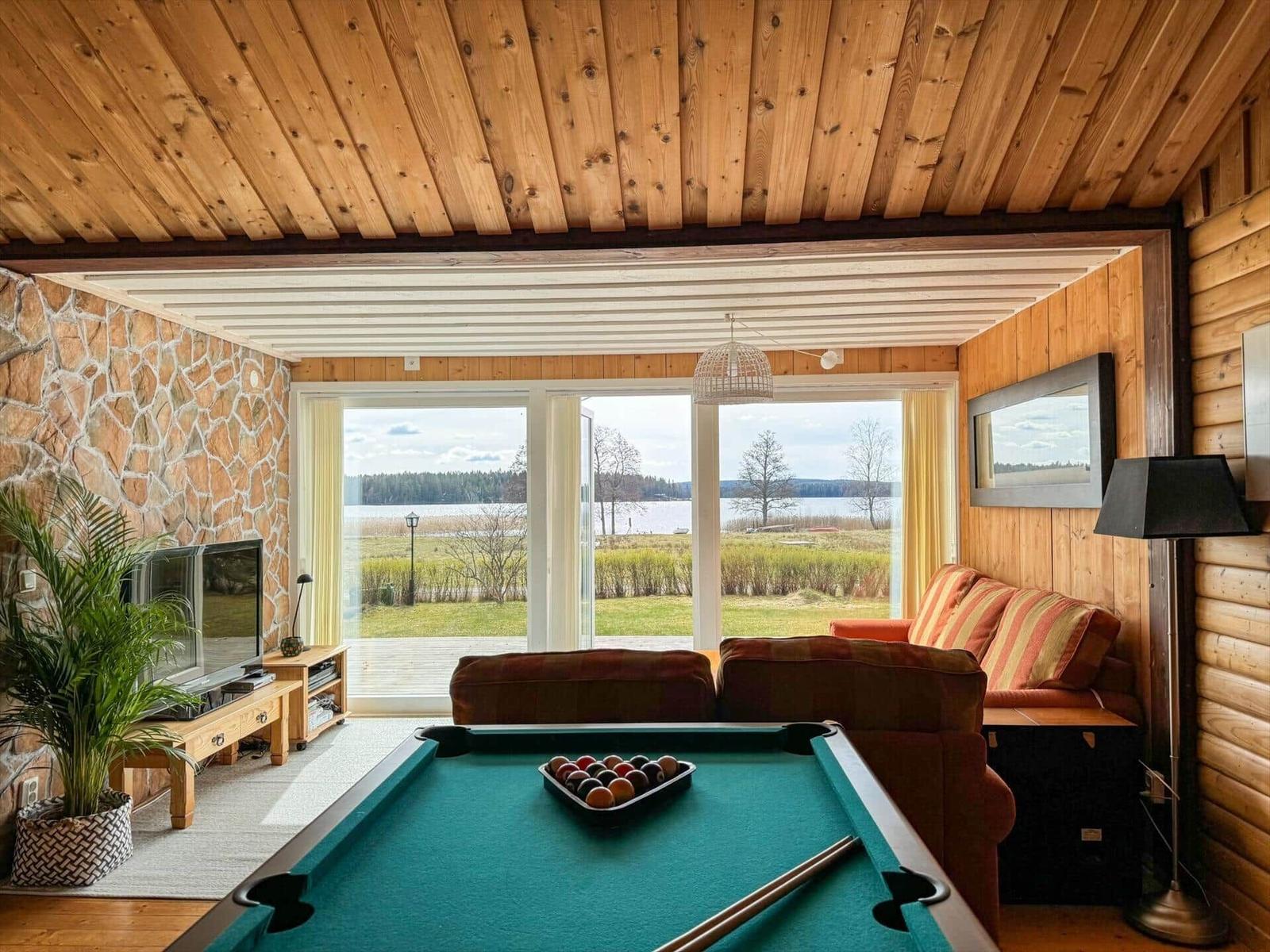 Ein Raum mit Billardtisch, Sofa und Fernseher. Durch große Fenster ist ein See zu sehen.