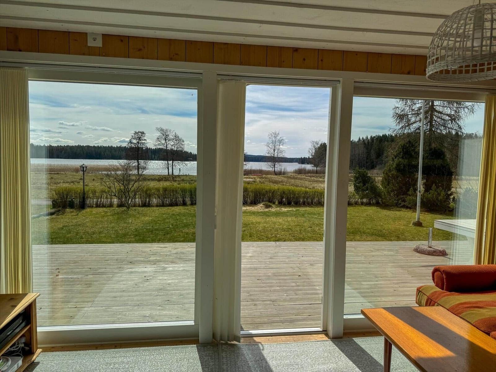 Große Glasfront mit Blick auf See und Grünfläche. Holzterrasse und Sitzgelegenheit.