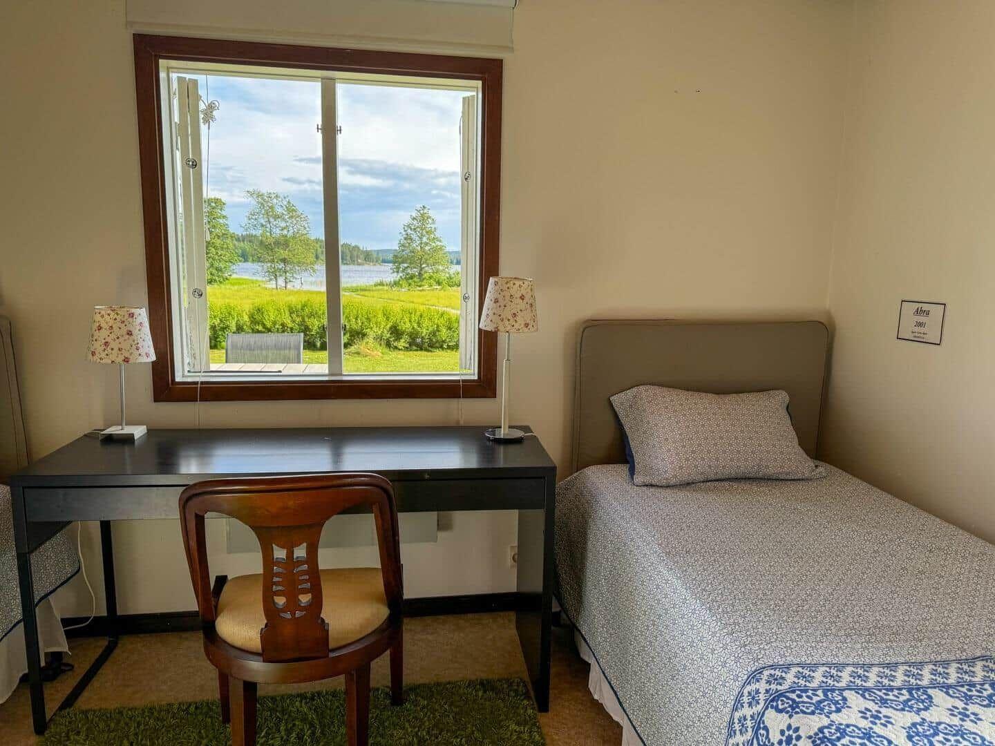 Ein Zimmer mit Bett, Schreibtisch und Stuhl. Durch das Fenster sieht man einen See.