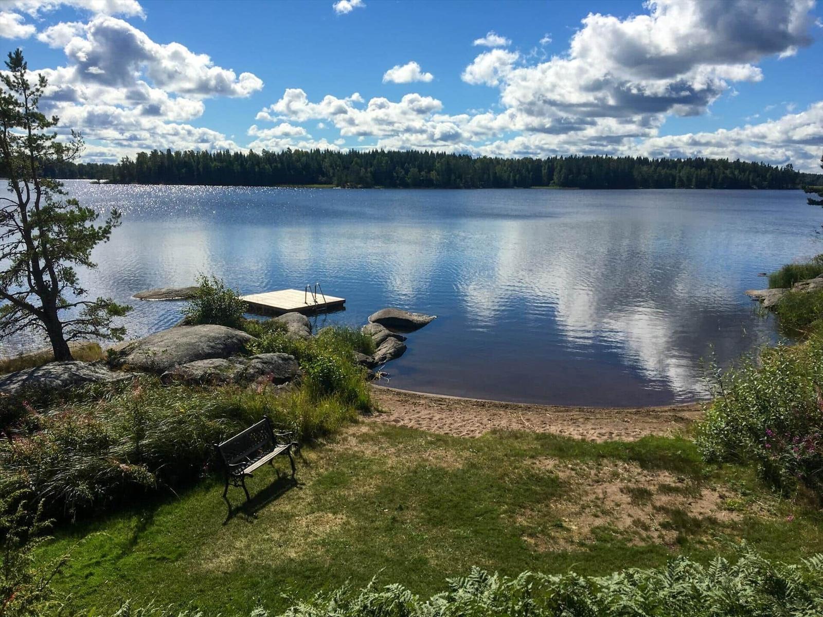 Lakeside with wooden dock, bench, and forested shore under cloudy sky.