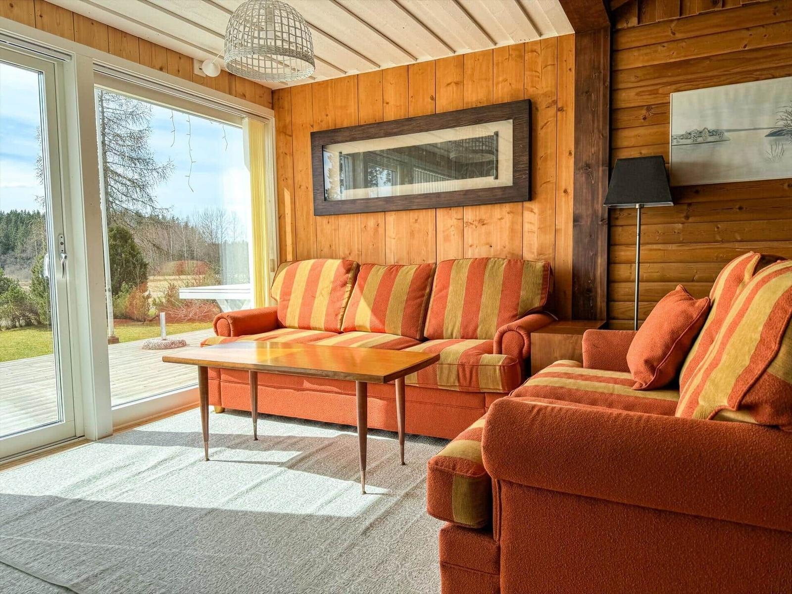 Wohnzimmer mit Holzwand, Sofa und Sessel. Große Glasfront zum Garten.