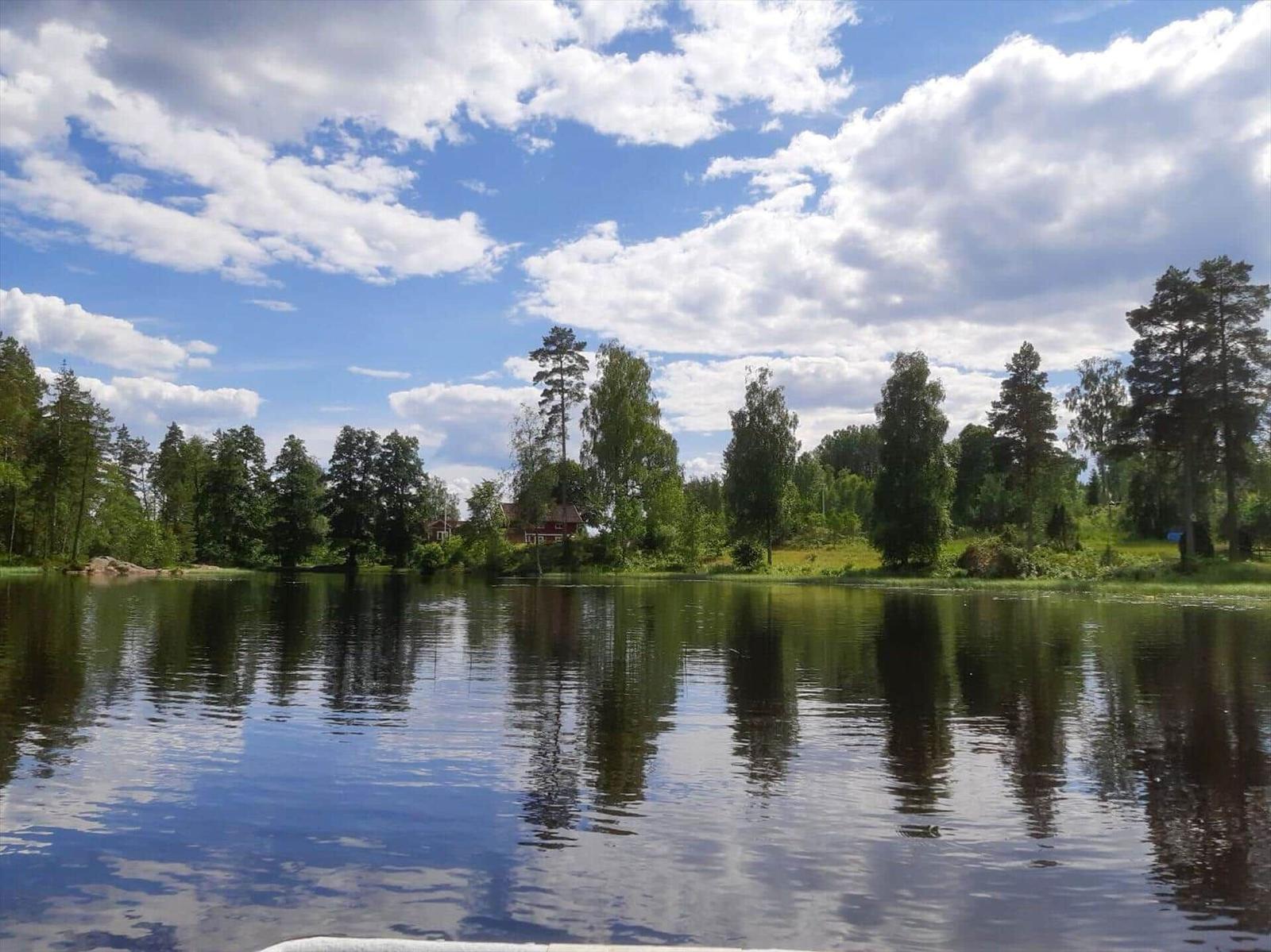 See mit Spiegelung, Bäume und roter Hütte am Ufer unter blauem Himmel mit Wolken.
