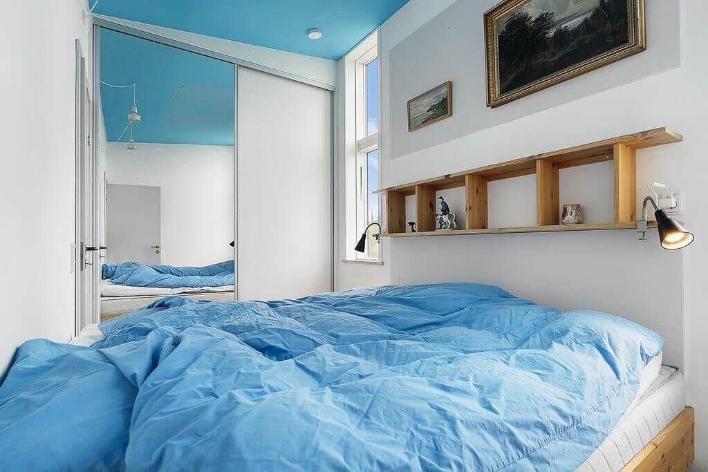 Schlafzimmer mit blauem Bett und Decke, Spiegeltür und Holzregal an der Wand.