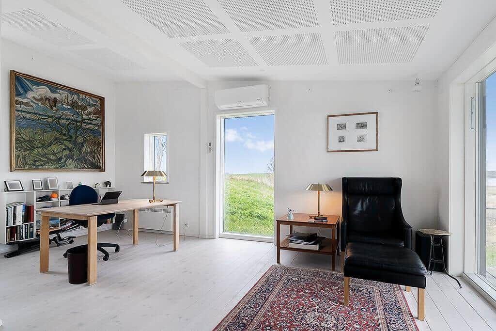 Ein Arbeitszimmer mit Schreibtisch, Stuhl und Sessel. Große Fenster bieten Blick auf Grünflächen.