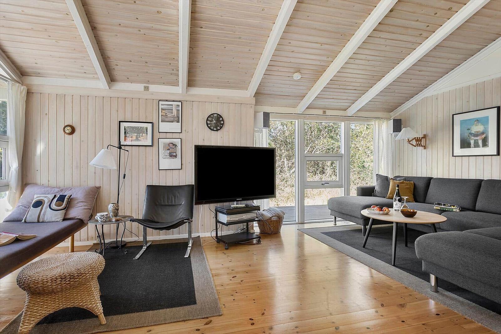 Wohnzimmer mit Sofa, Fernseher und Holzboden. Große Fenster und hölzerner Deckenrahmen.