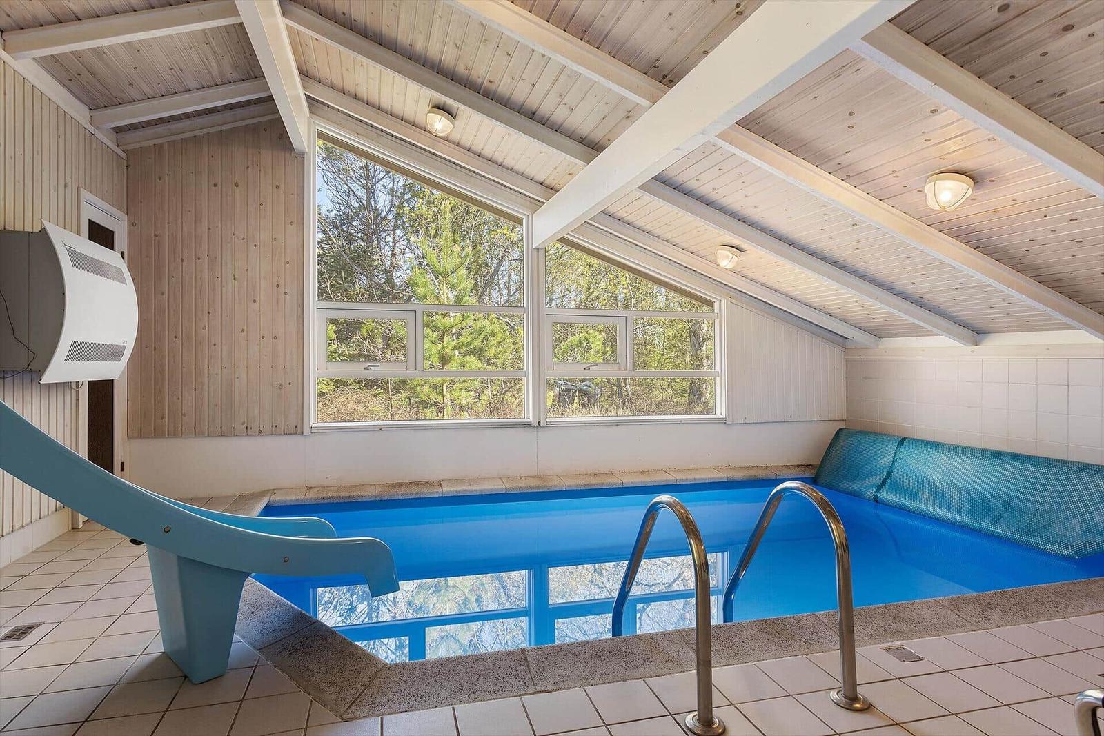 Innere Schwimmhalle mit blauem Pool, Holzdecke und Fenster zur Natur.