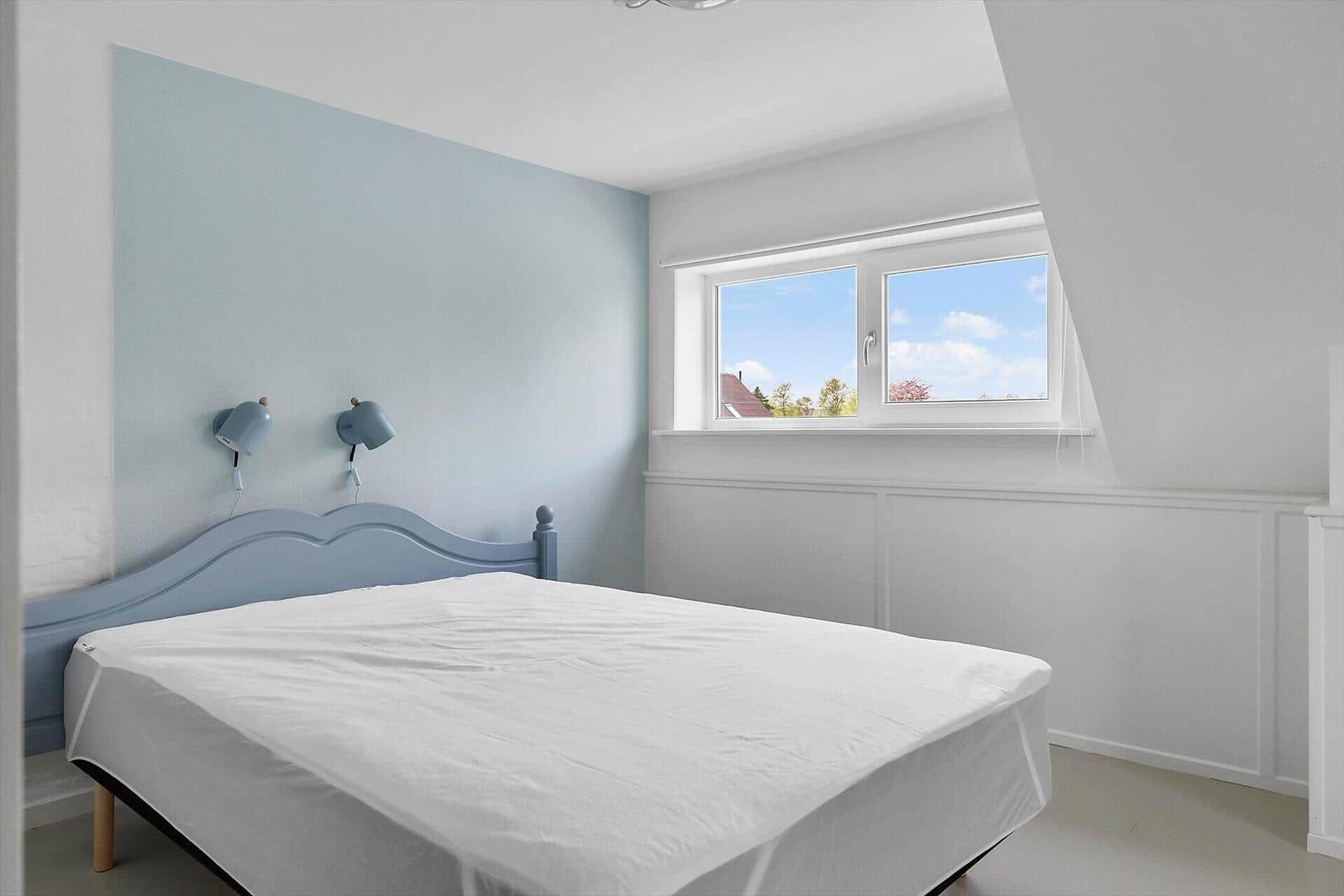 Schlafzimmer mit Bett, Wandlampe und Fenster mit Blick auf Außenbereich.
