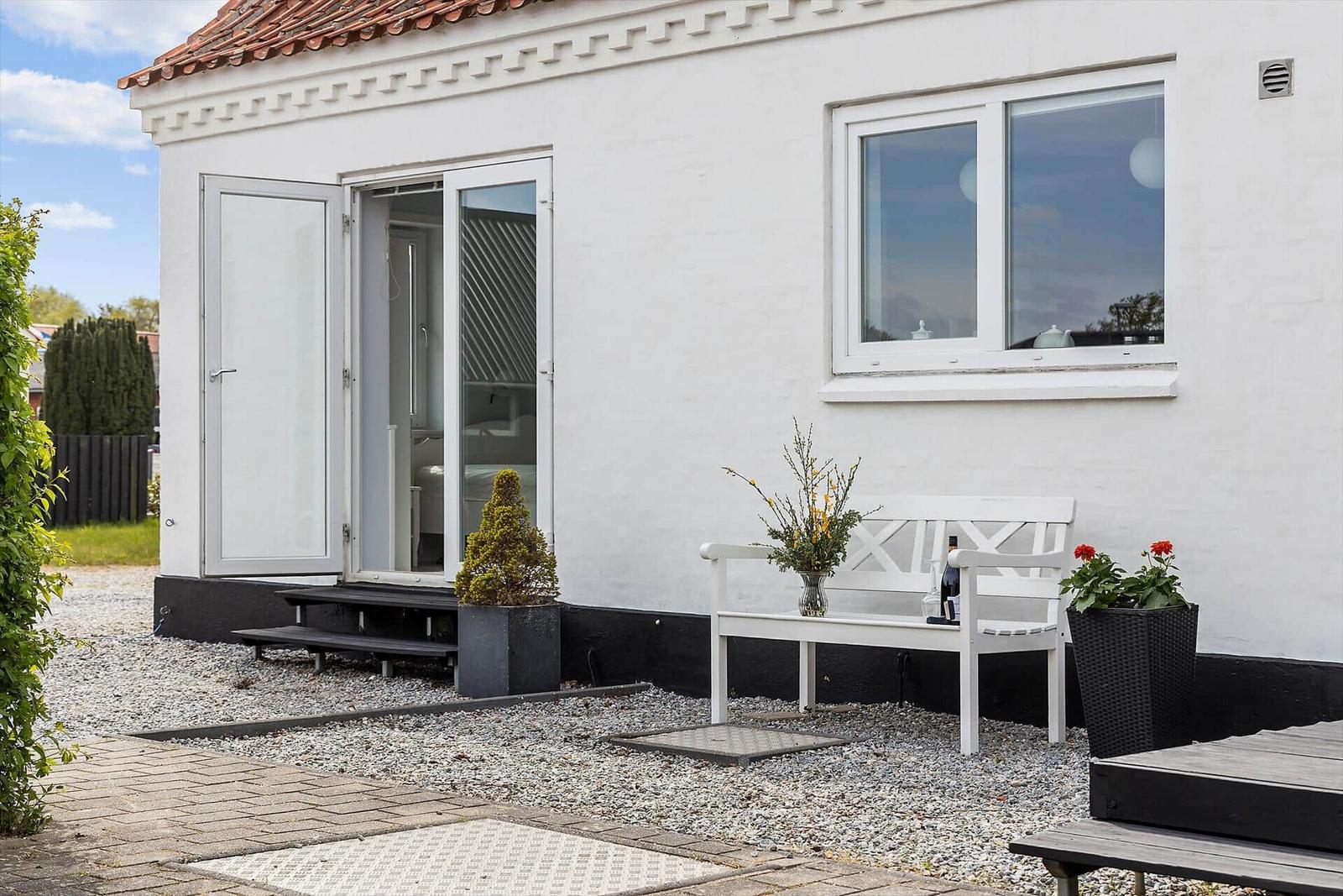 Weißes Haus mit Terrasse, Bank und Pflanzen. Glastür führt ins Innere.