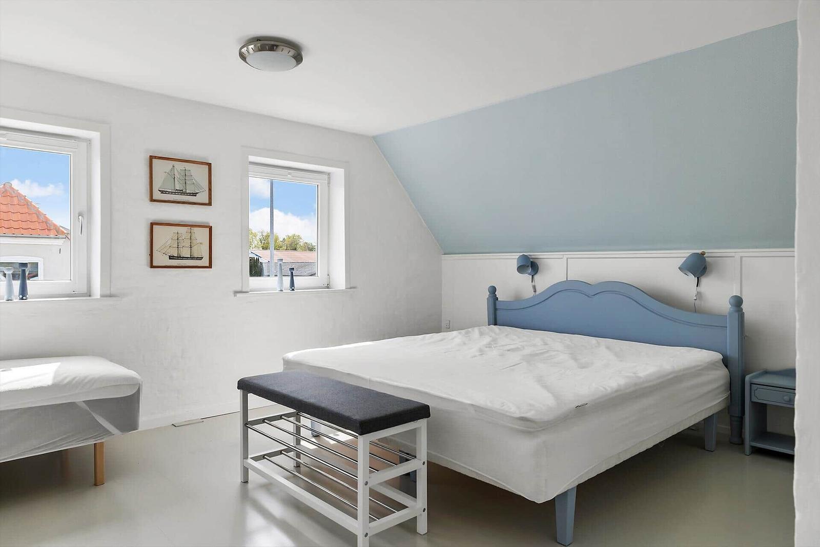 Schlafzimmer mit blauem Bett, zwei Fenstern und Wandbildern von Schiffen.