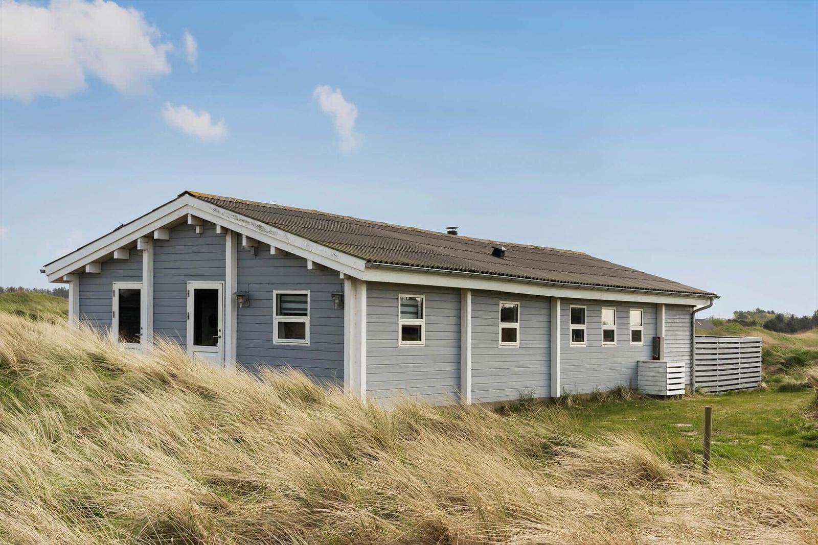 A gray holiday home with white window frames and roof gutter, surrounded by tall grass.