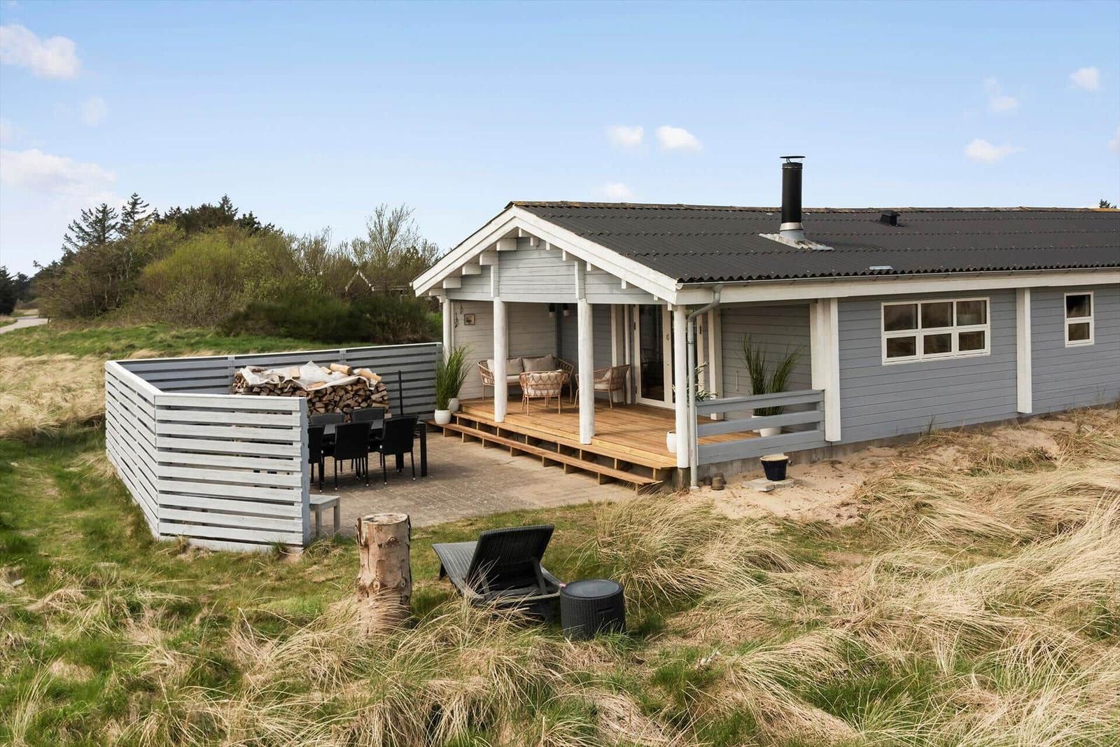 A gray house with a terrace, garden furniture, and a woodpile. Surrounded by grass and trees.