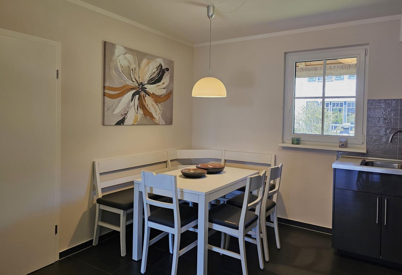 Essbereich mit weißem Tisch, Stühlen und Wandbild. Fenster und Küchenbereich sichtbar.