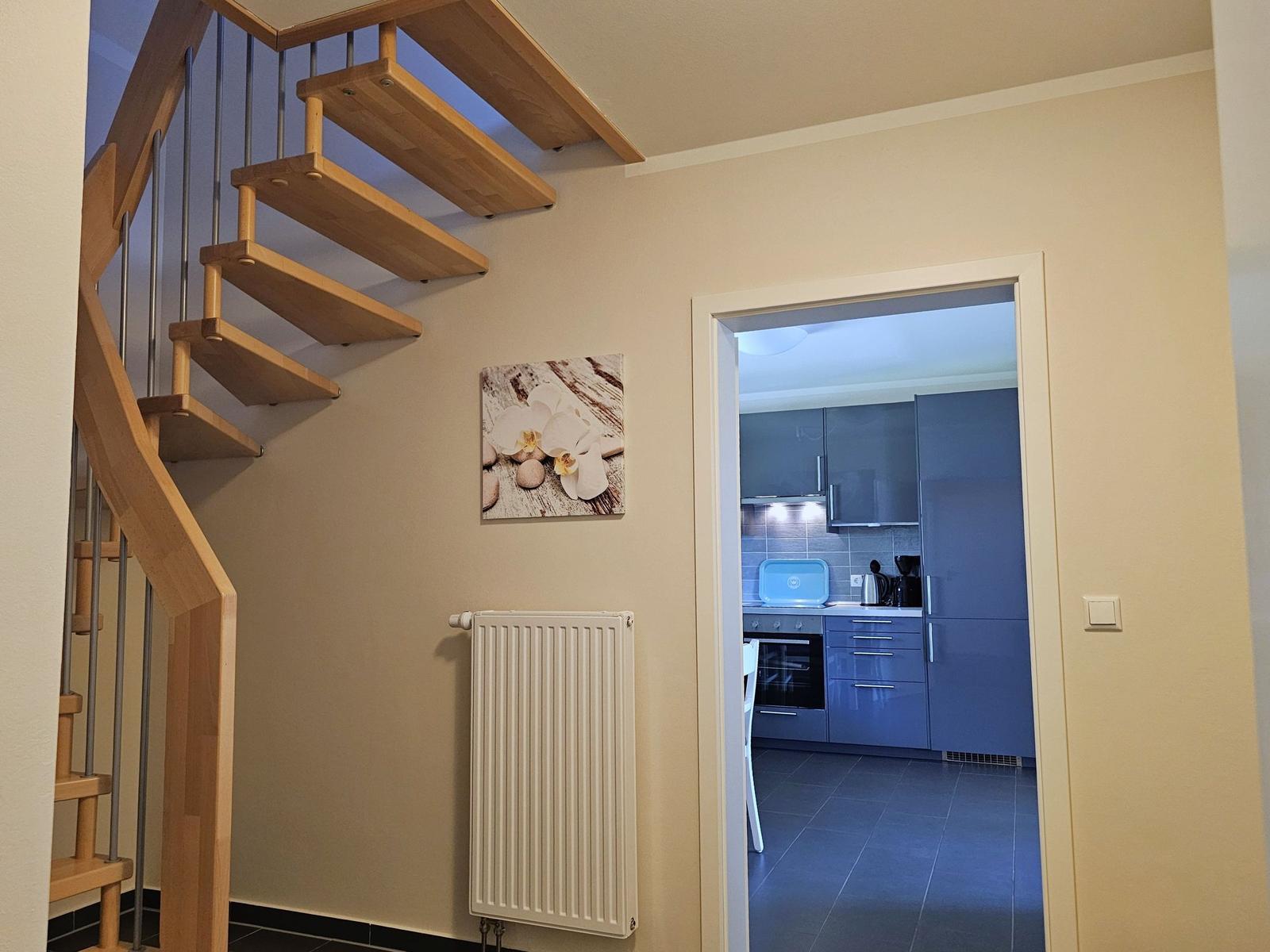 Holztreppe führt zur oberen Etage. Küche mit blauen Schränken ist sichtbar.