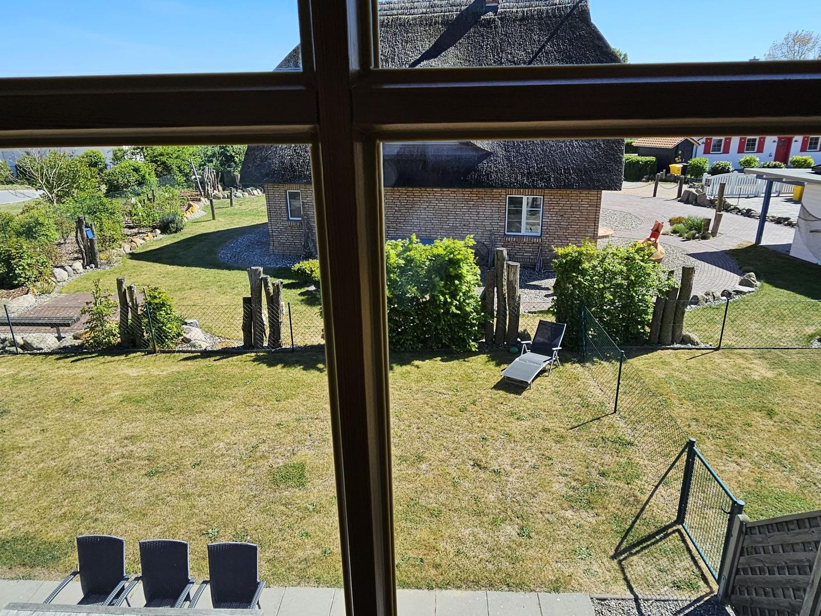 Von innen betrachtet: Garten mit Liegestuhl und Blick auf ein Haus mit Strohdach.