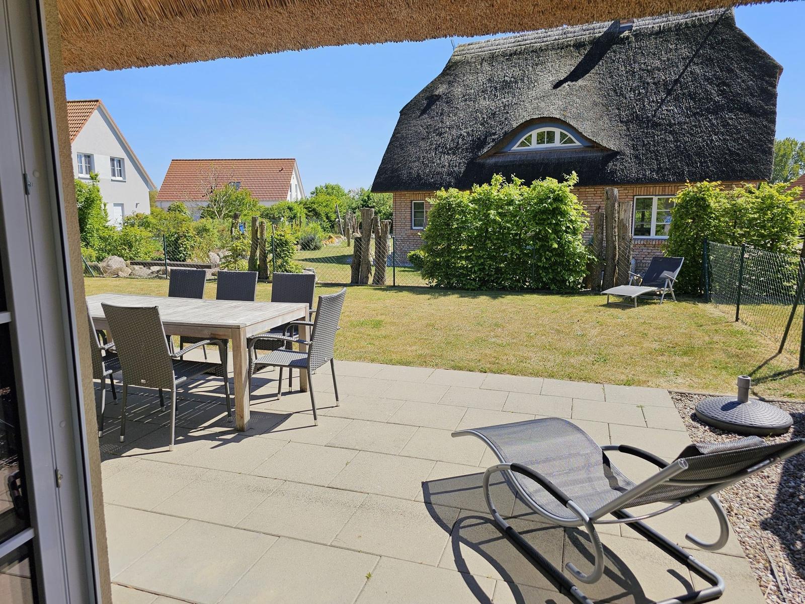 Terrasse mit Tisch, Stühlen und Liegestuhl. Blick auf Garten und strohgedecktes Haus.
