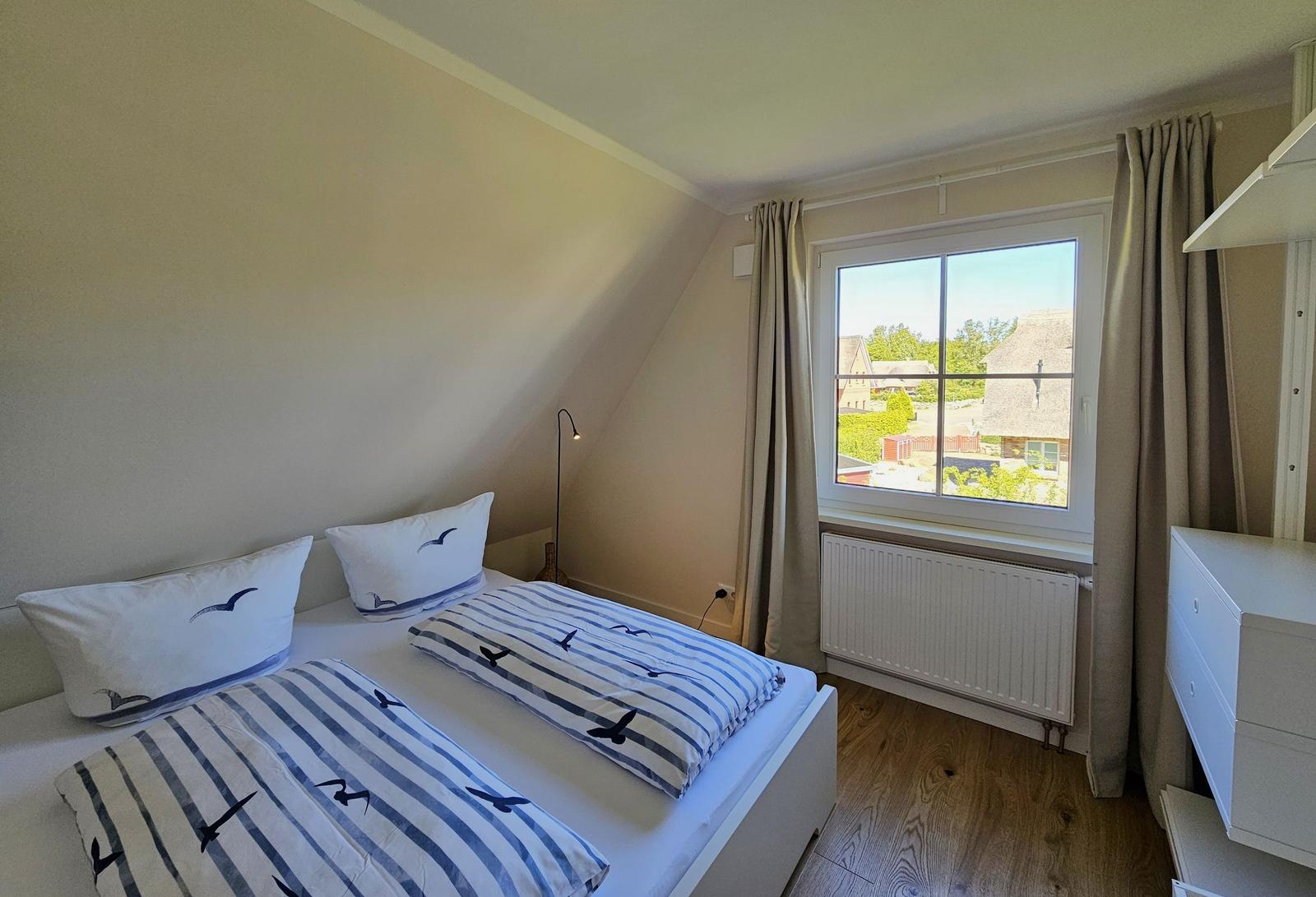 Doppelbett mit Vogelmotiv, Fenster mit Blick auf Dächer und Bäume, Holzboden, weißer Schrank und Heizkörper.