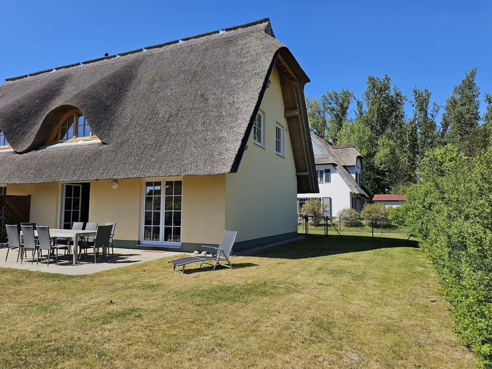 Haus mit Strohdach, Terrasse mit Tisch und Stühlen, Garten und Liegestuhl.
