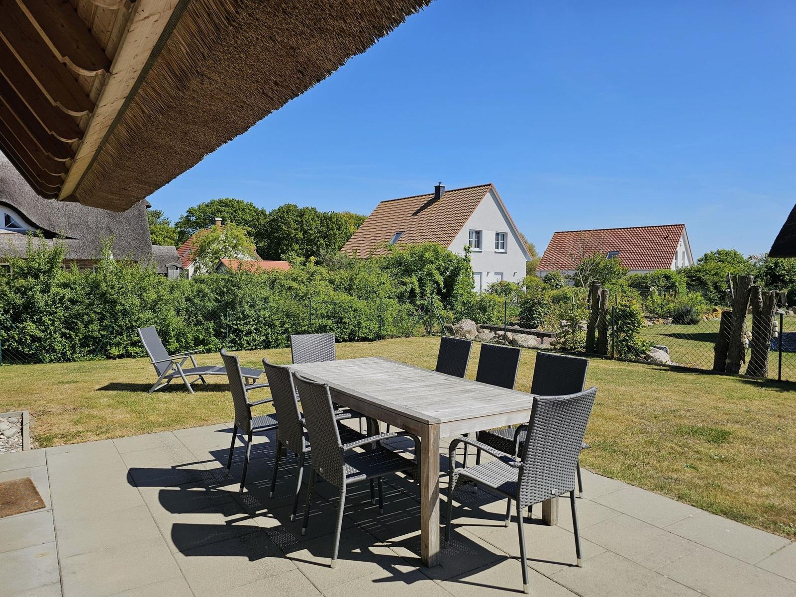 Terrasse mit Esstisch und Stühlen unter strohgedecktem Dach. Grünfläche und Häuser im Hintergrund.