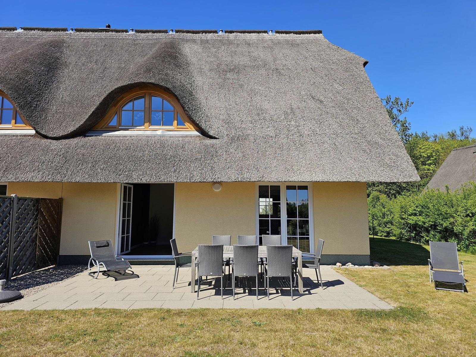 Haus mit Strohdach, Terrasse mit Tisch und Stühlen, Garten und Sitzgelegenheiten.
