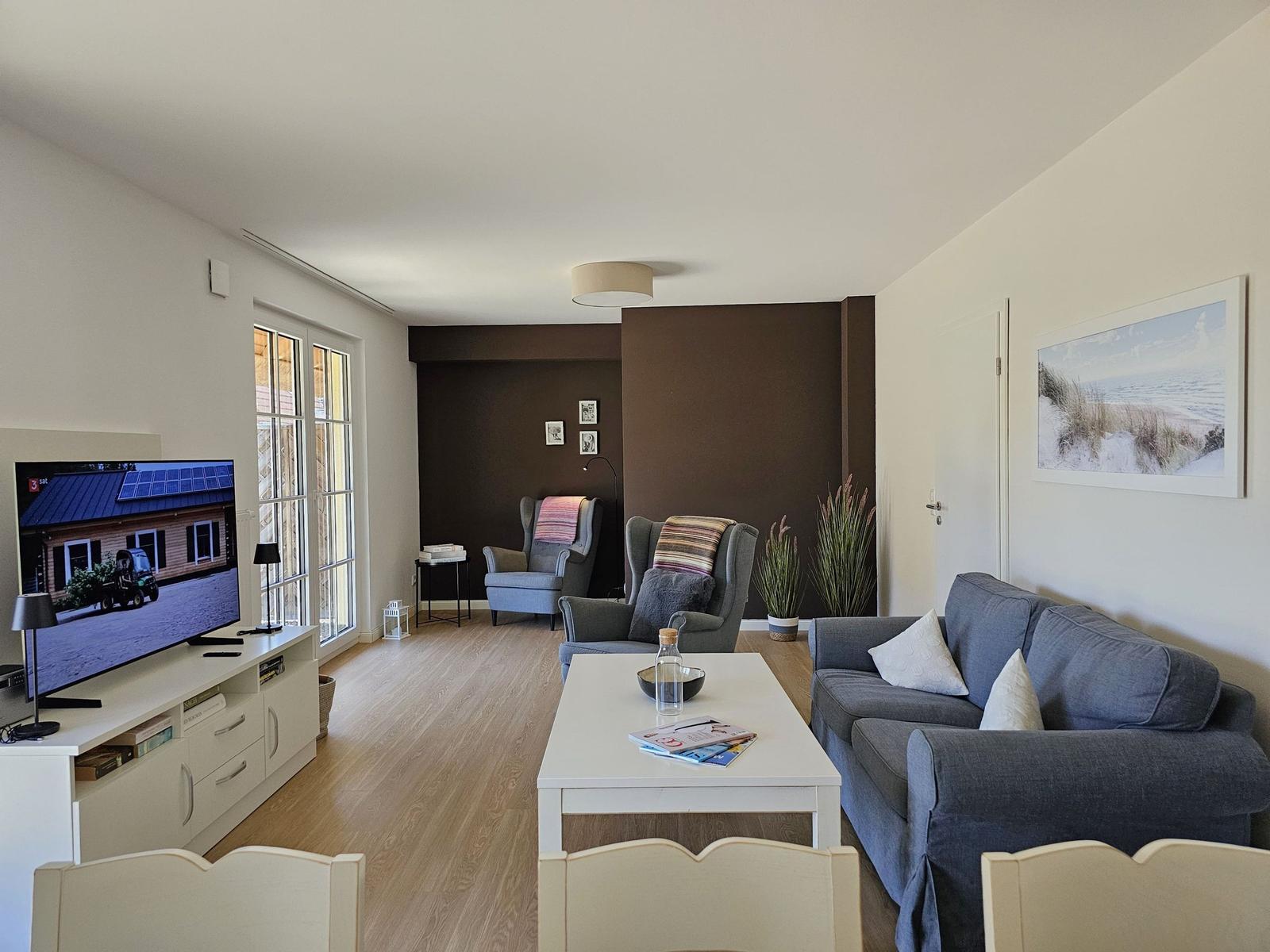 Wohnzimmer mit Sofa, Sesseln und Fernseher. Holzfußboden und große Fenster.