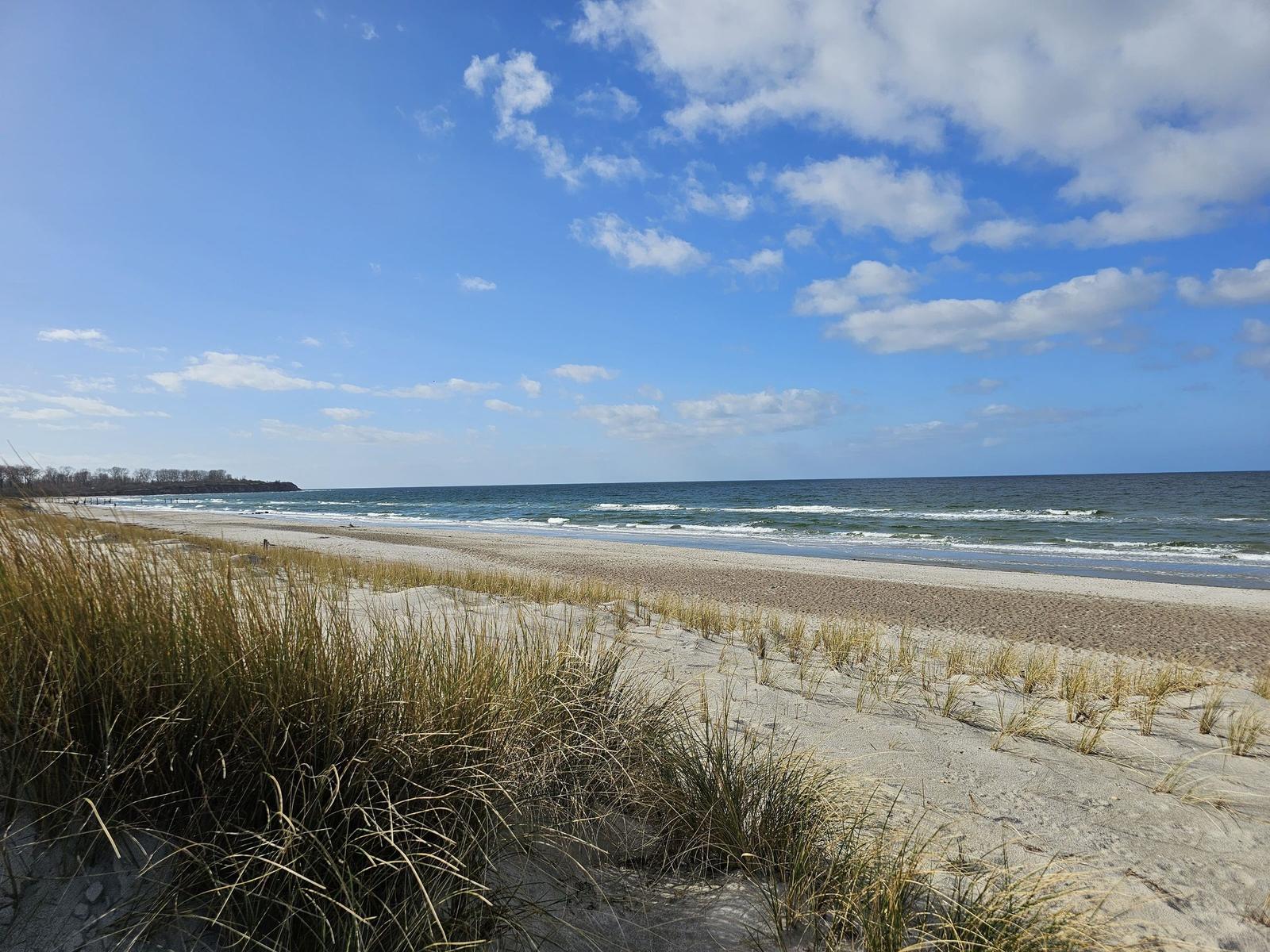 Strand mit Dünen und Wellen unter blauem Himmel mit weißen Wolken.
