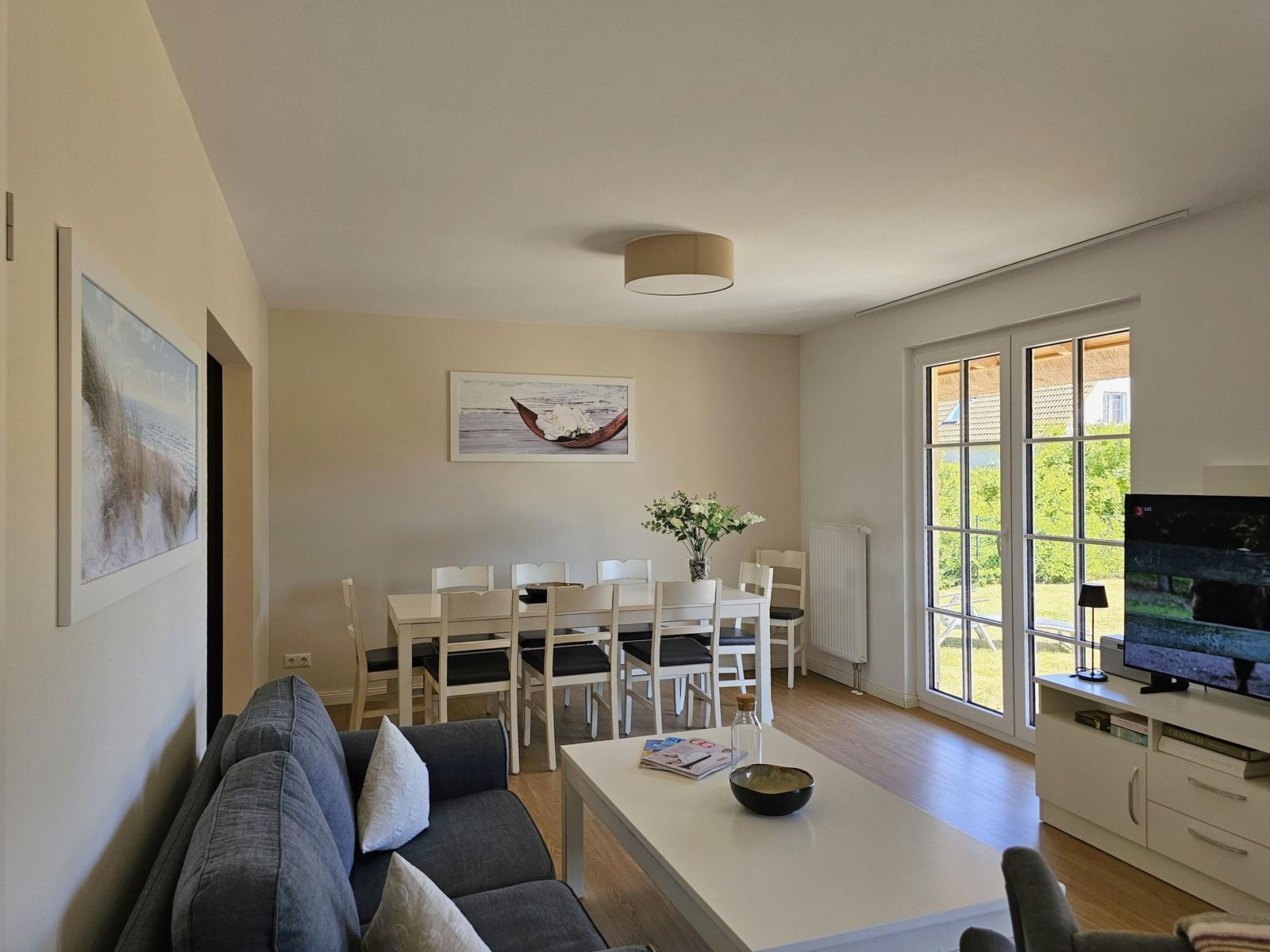 Wohn- und Essbereich mit Sofa, Tisch und Stühlen, Fernseher und Blick auf Garten.