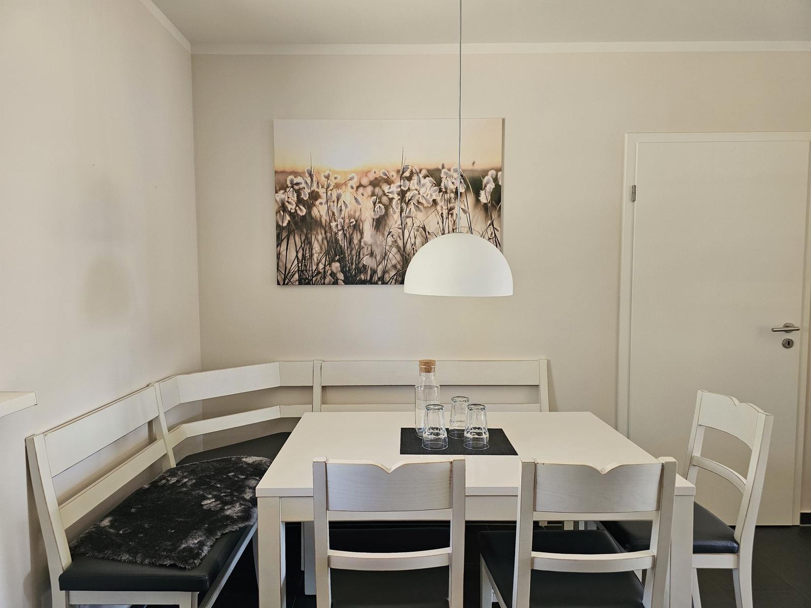 Essbereich mit weißem Tisch, Bänken und Stühlen. Wandbild und Pendelleuchte.