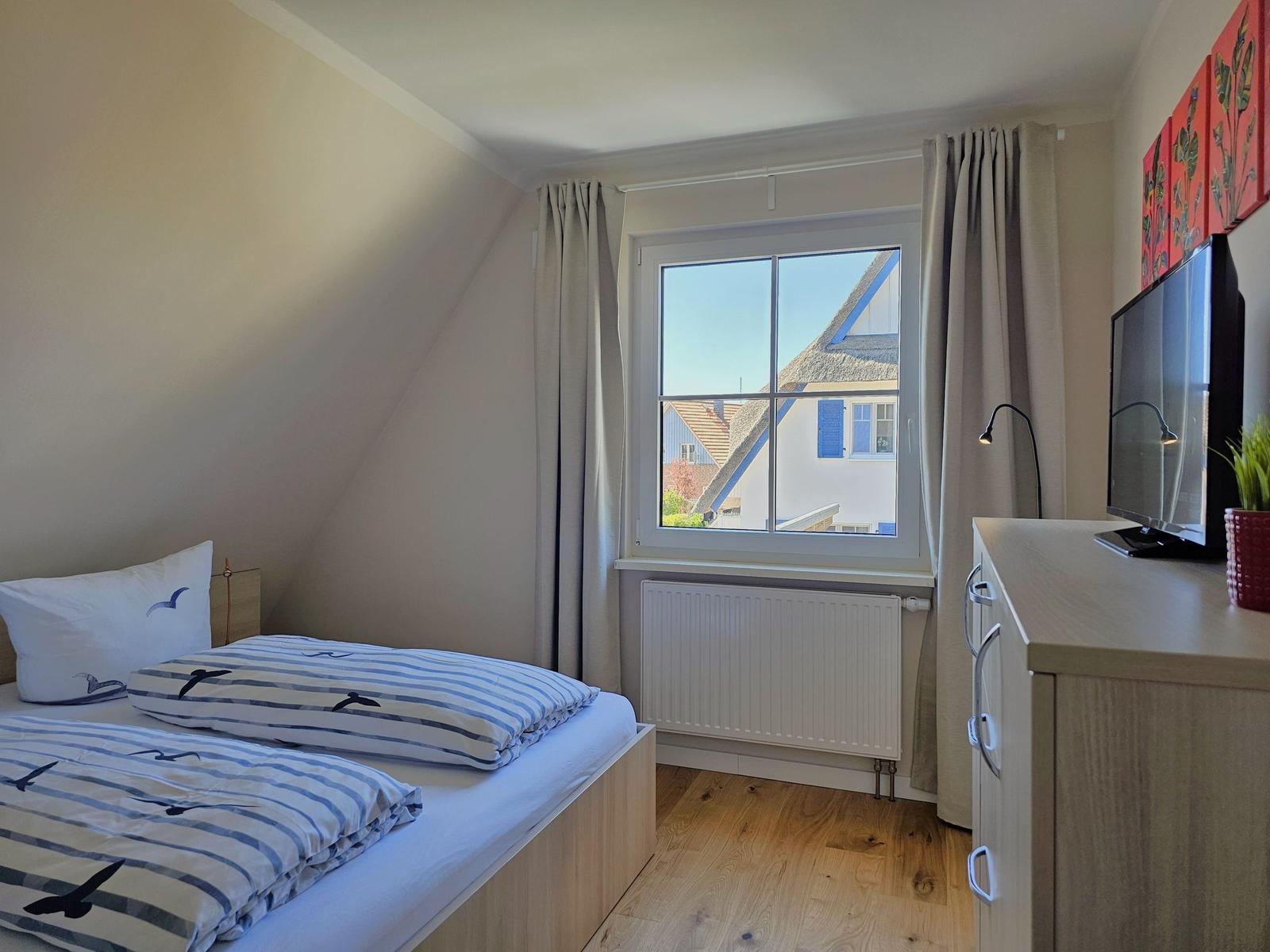 Schlafzimmer mit Bett, Fenster und Schrank. Blick auf Dachhäuser.