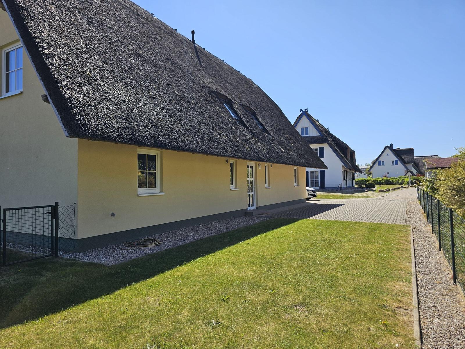 Haus mit Strohdach, Garten und gepflastertem Weg. Weitere Häuser im Hintergrund.