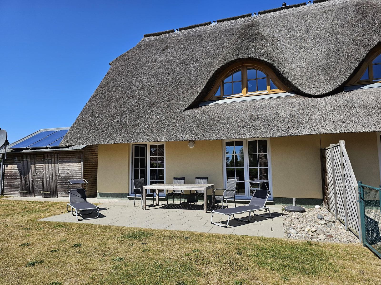 Haus mit Strohdach, Terrasse mit Tisch, Stühlen und Liegestühlen.