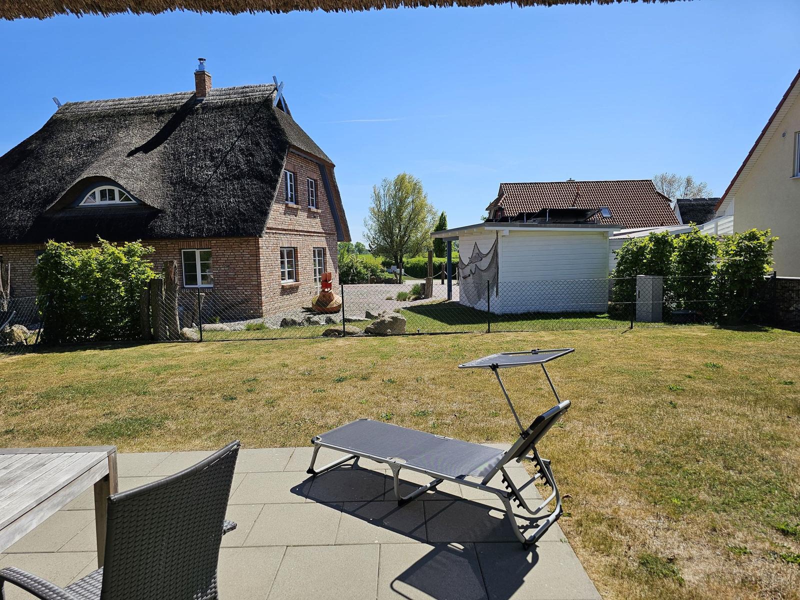 Terrasse mit Liegestuhl und Tisch. Hintergrund: Haus mit Strohdach und Garten.