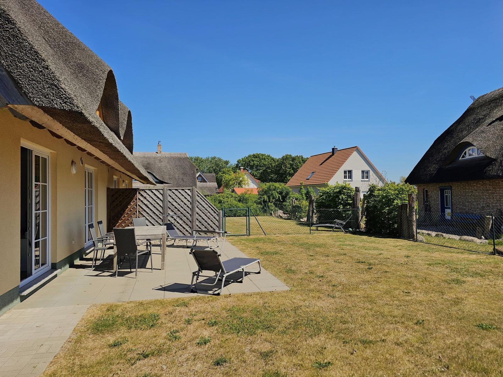 Terrasse mit Tisch, Stühlen und Liegen. Hinterhof mit Grasfläche und Nachbarhäusern.