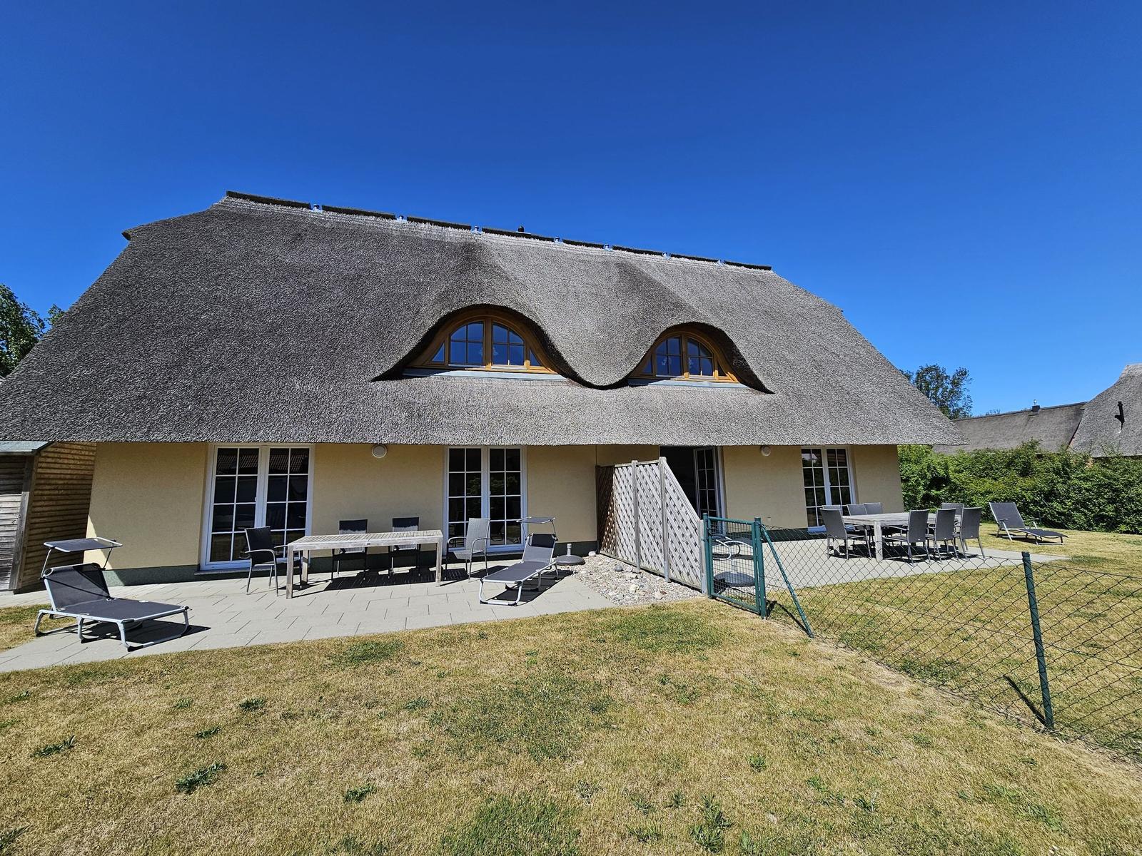 Haus mit Strohdach, Terrasse und Garten mit Liegestühlen und Tisch.