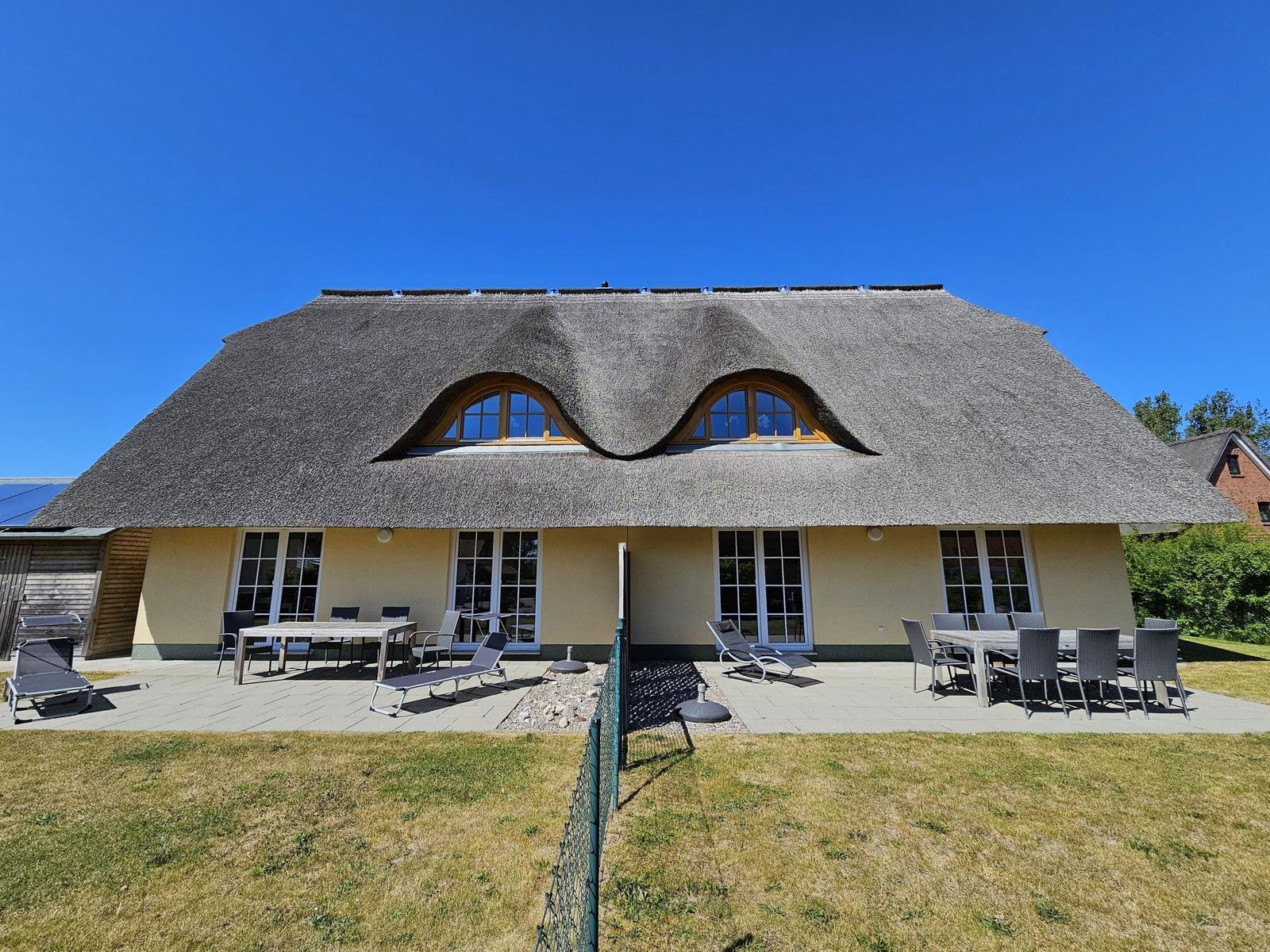 Haus mit Strohdach und großem Gartenbereich mit Tischen und Liegen.