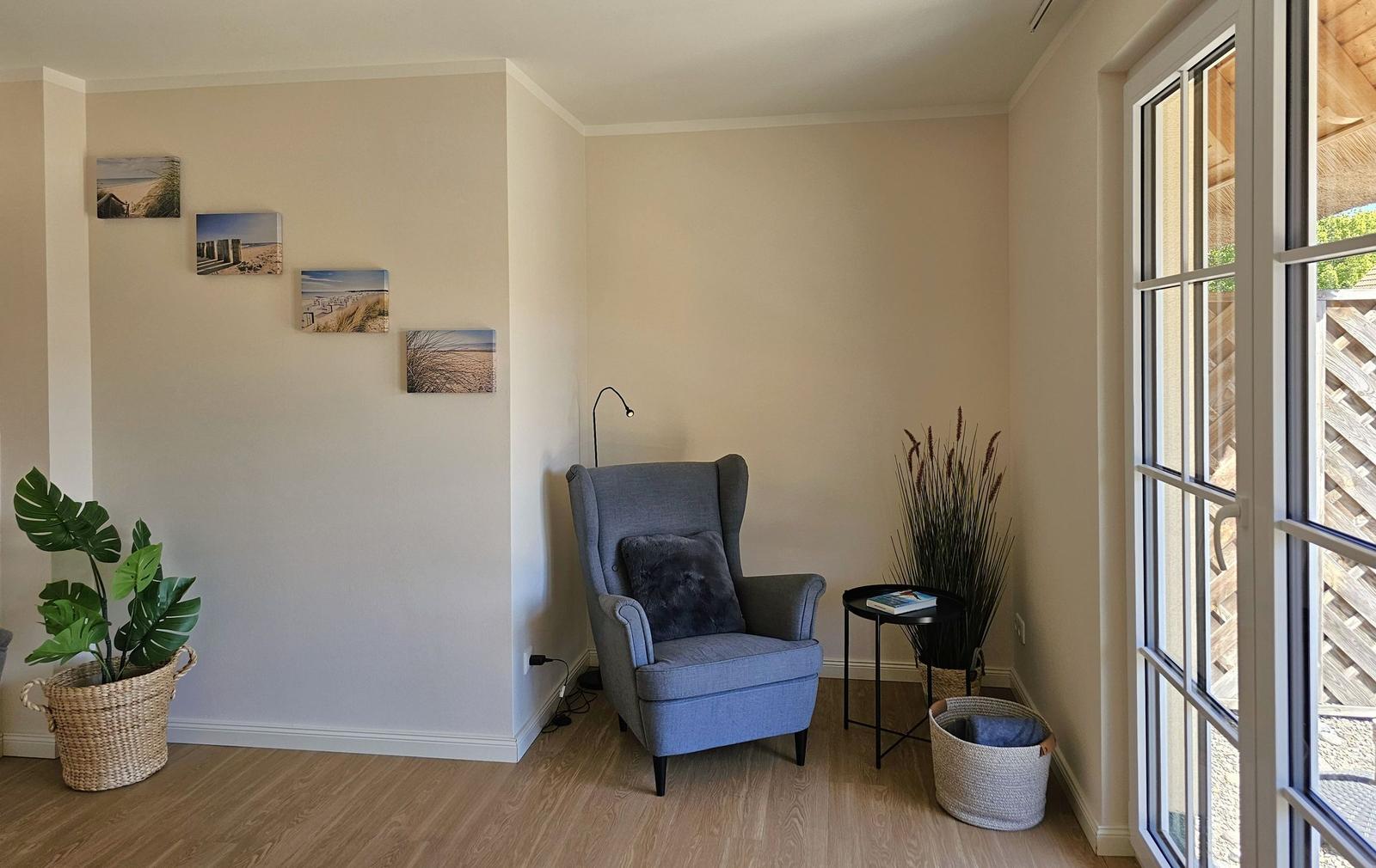 Ein Raum mit grauem Sessel, Pflanzen und Wandbildern an der Wand.