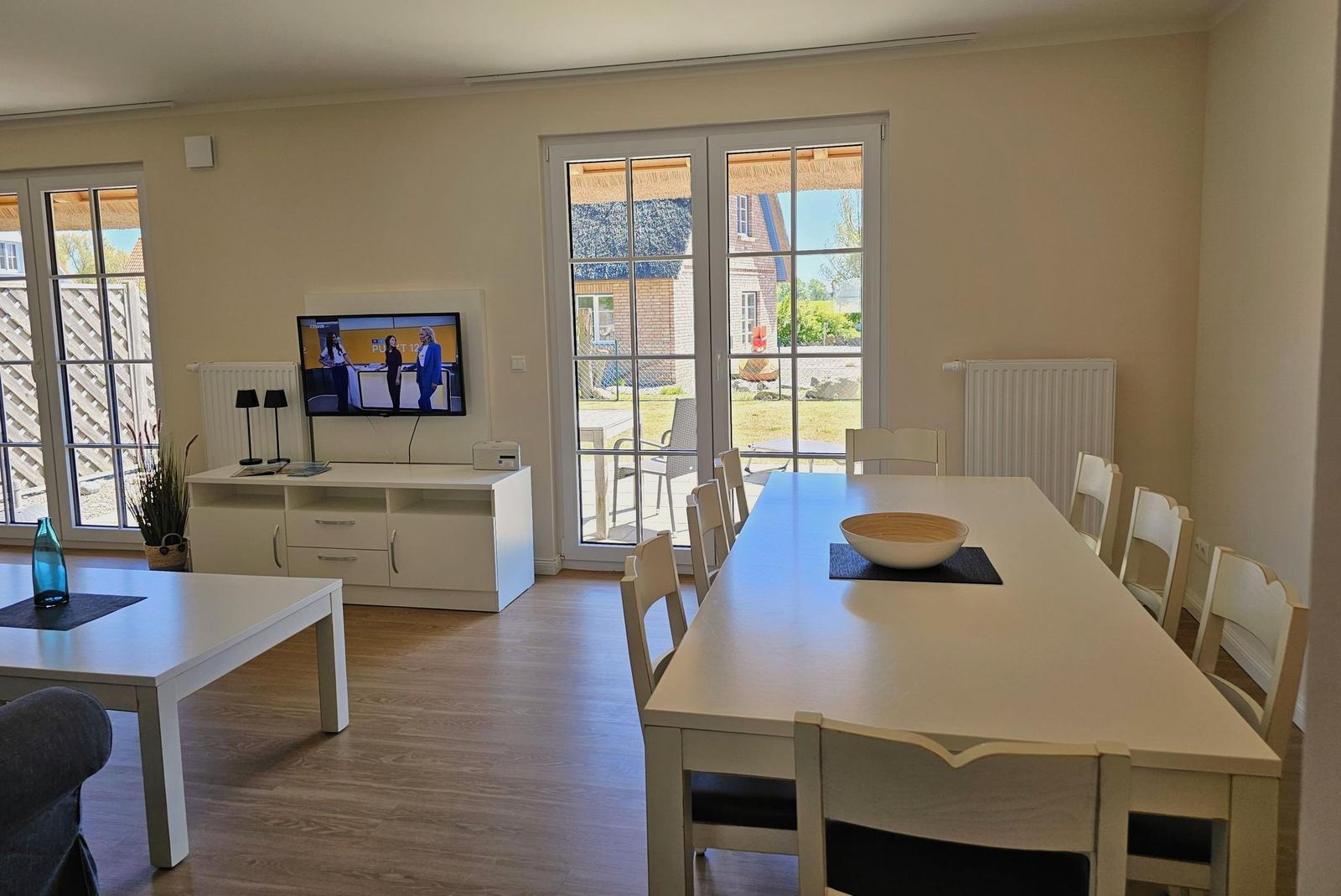 Essbereich mit langem Tisch und Stühlen. Fernseher an Wand. Terrasse durch Glastüren erreichbar.
