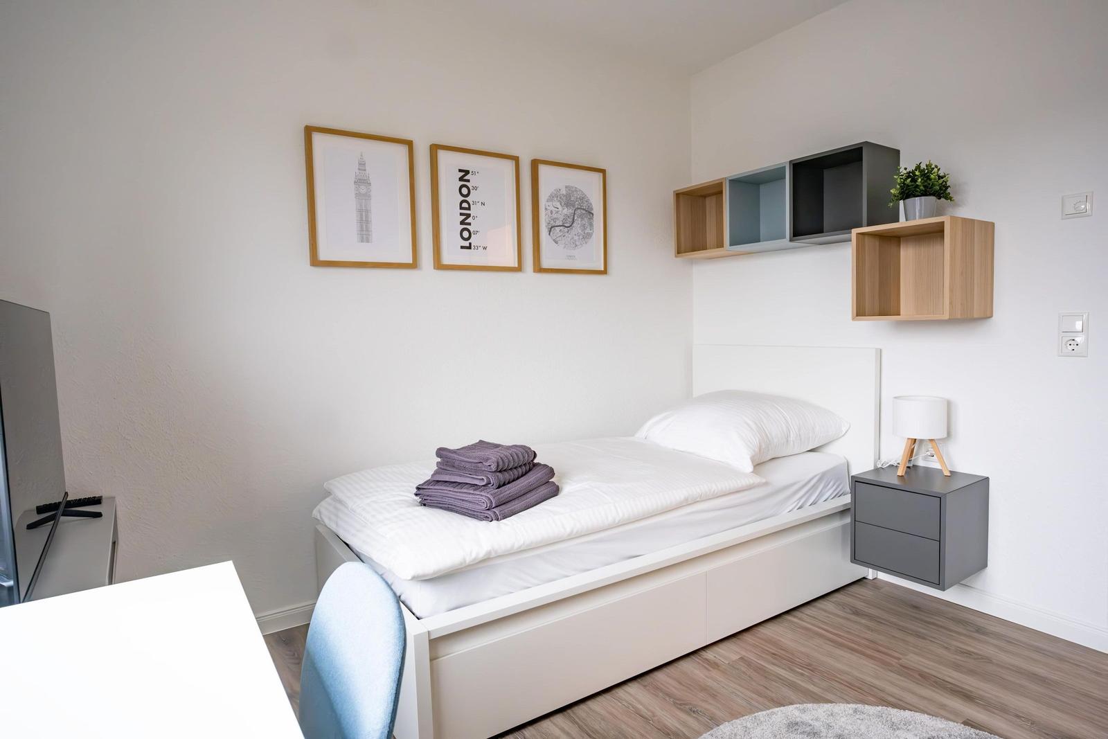 Schlafzimmer mit Bett, Nachttisch und Wandregalen. An der Wand hängen drei Bilder.