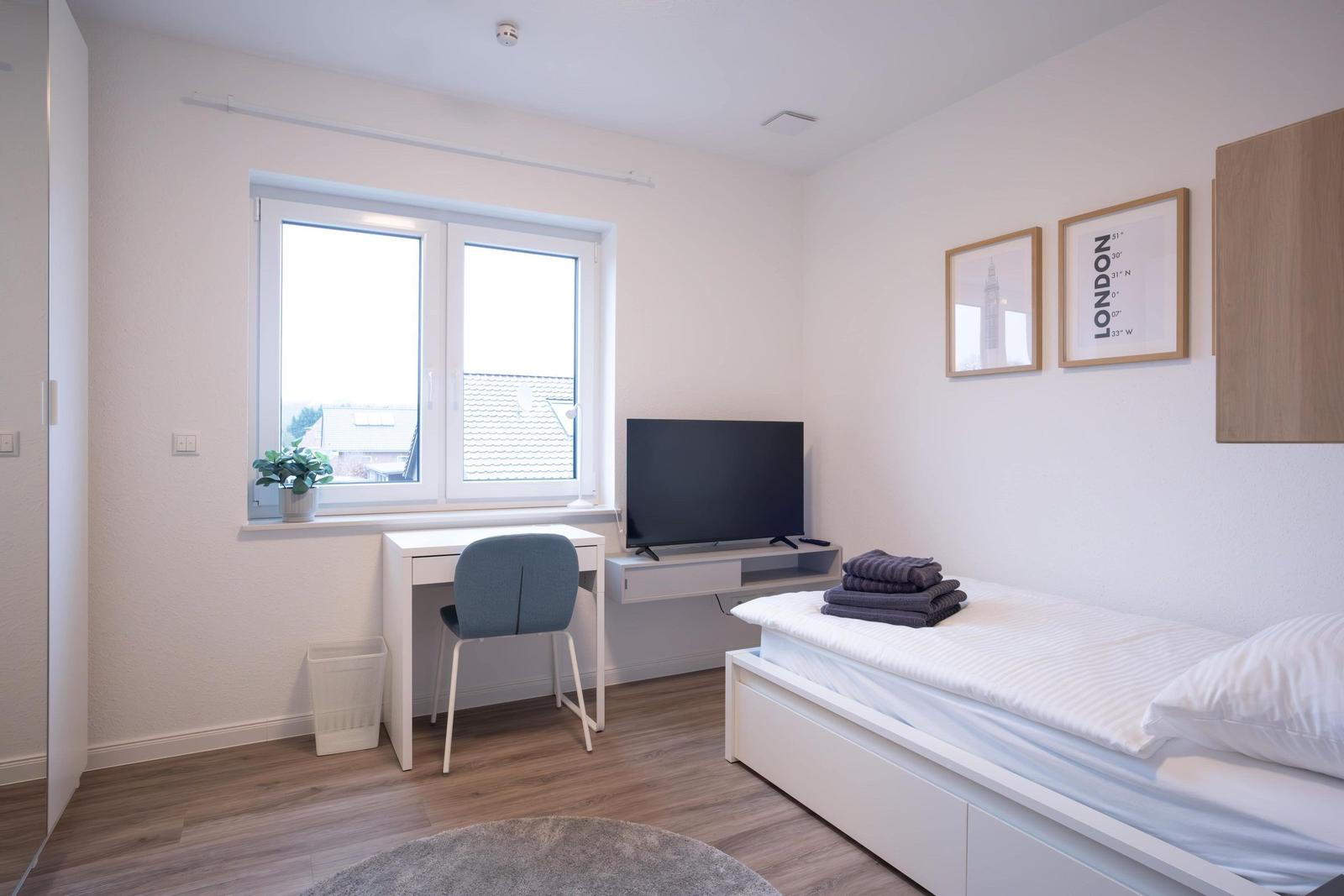 Schlafzimmer mit Bett, Schreibtisch, TV und Fenster.