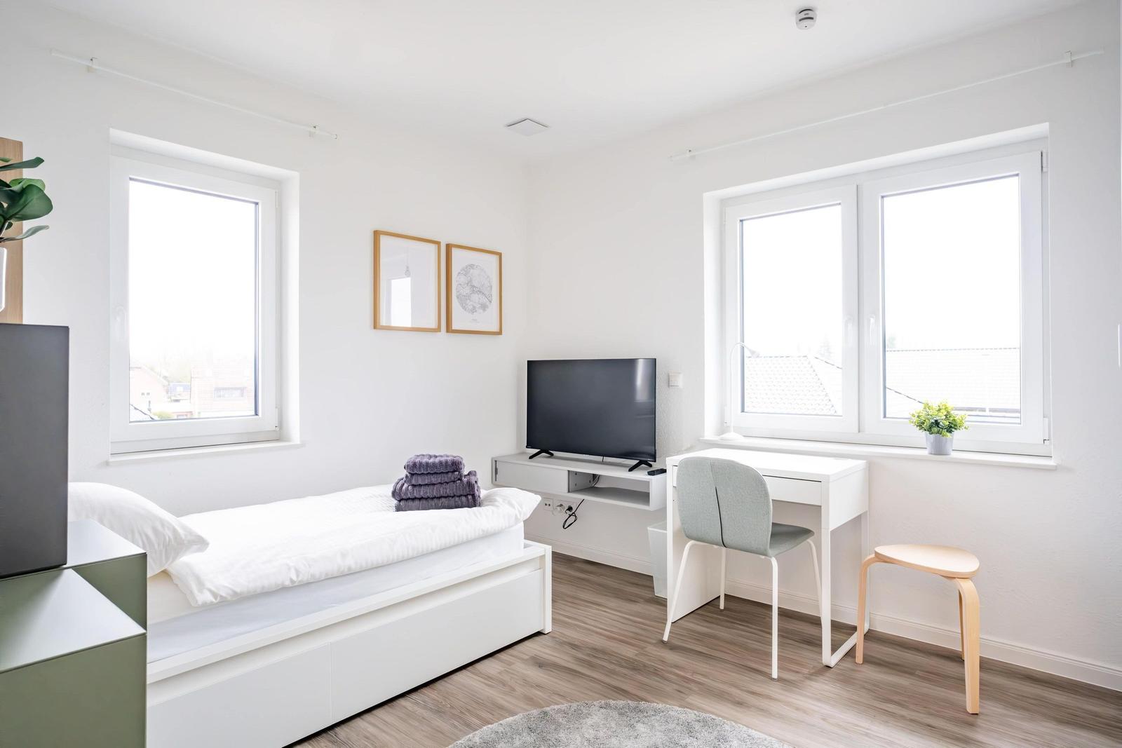 Schlafzimmer mit Bett, Fernseher, Schreibtisch und Fenstern.