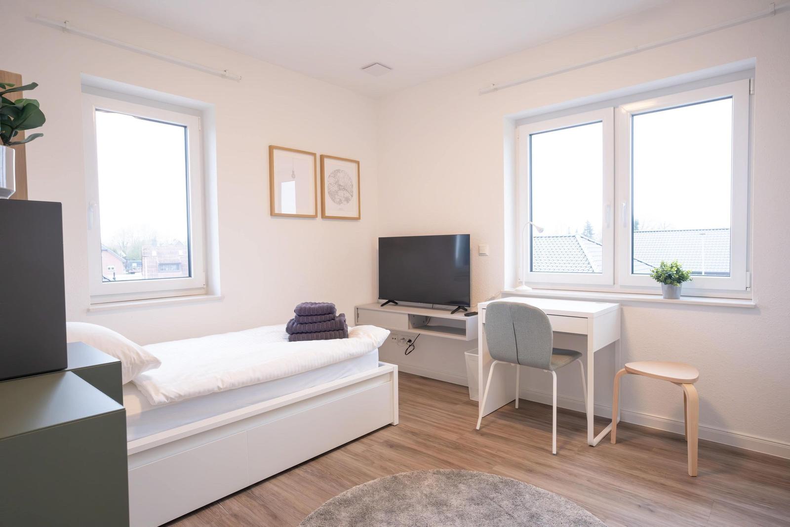 Schlafzimmer mit Bett, Schreibtisch, Fernseher und zwei Fenstern.