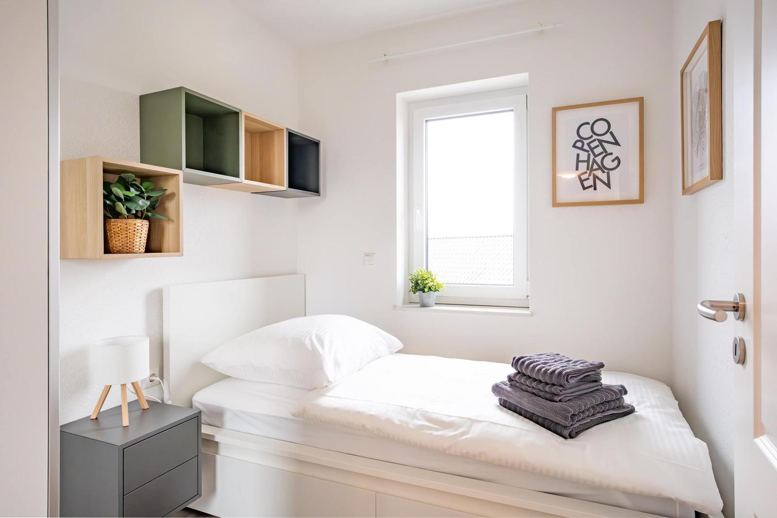 Schlafzimmer mit Bett, Nachttisch und Wandregalen. Fenster mit Pflanze und Bild.