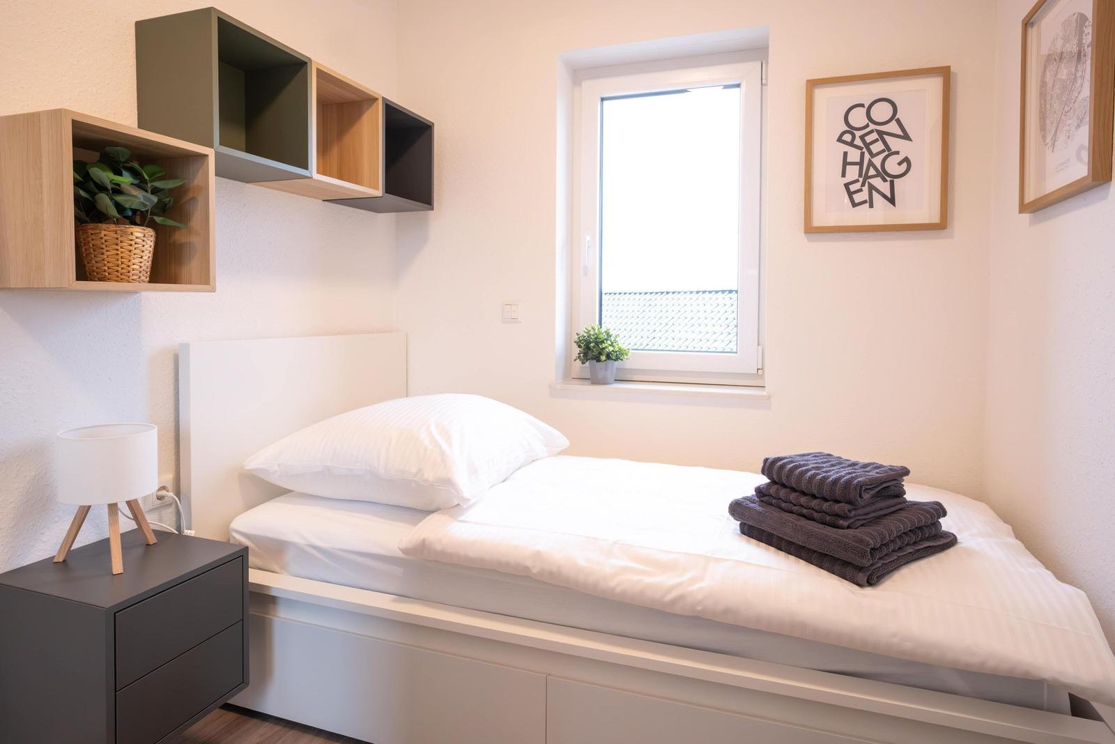 Schlafzimmer mit Bett, Nachttisch und Wandregal. Fenster und Bilder an der Wand.