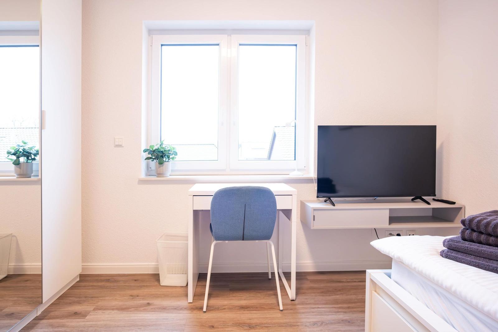 Schlafzimmer mit Schreibtisch, TV und Fenster. Bett mit Handtüchern. Holzboden und weiße Wände.
