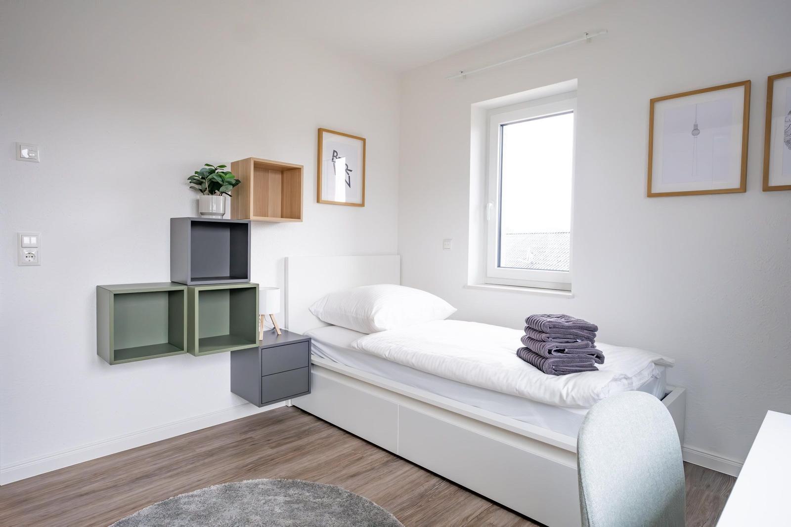 Ein Schlafzimmer mit weißem Bett, Holzboden und Wandregalen. An der Wand hängen Bilder.