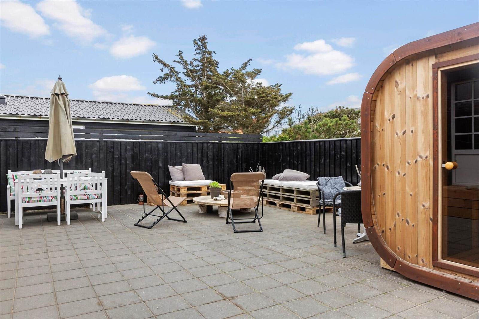 Terrasse mit Sitzgelegenheiten, Sonnenschutz und Holzsauna. Hinterhof mit Zaun und Baum.