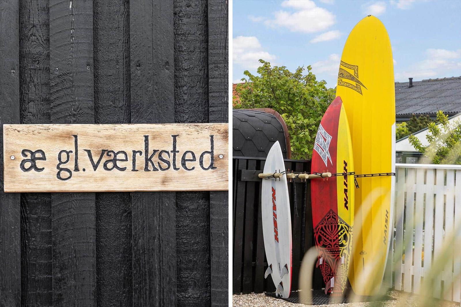 Holzschild mit Schrift an schwarzer Holzwand. Drei Surfbretter an Zaun im Garten.