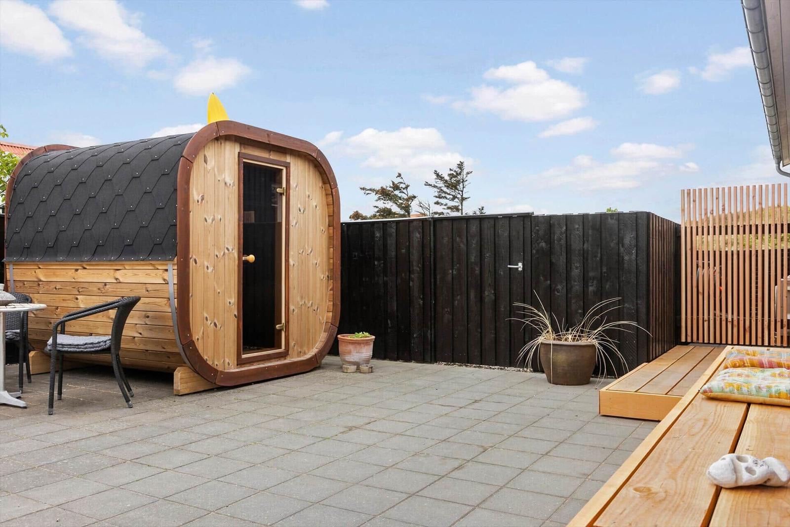 Außenbereich mit Holzsauna, Terrasse und Pflanzen. Hintergrund: Zaun und Bäume.