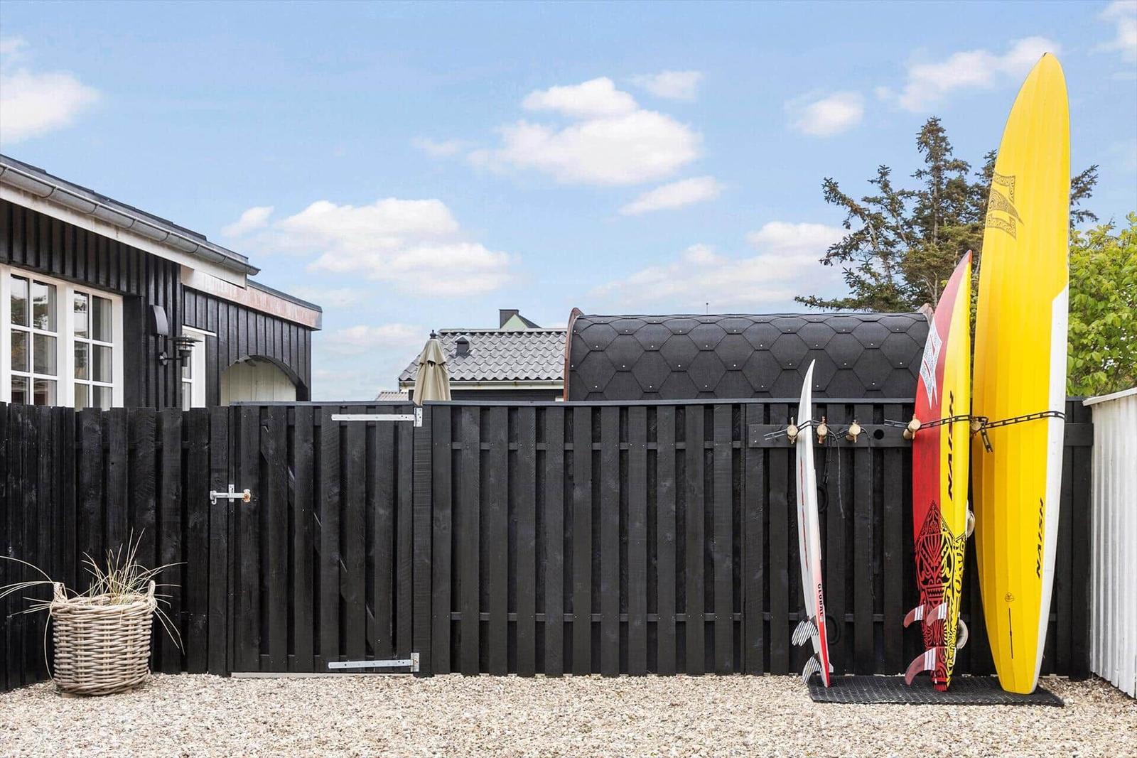Schwarzer Holzzaun mit drei Surfbrettern und einem Korb vor einem Haus.