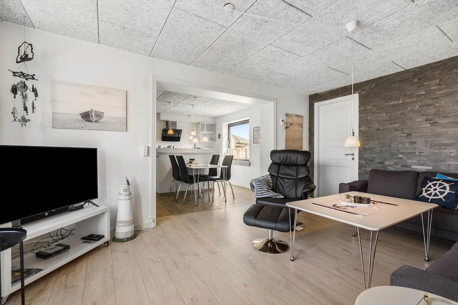 Wohnzimmer mit Sofa, Sessel und Fernseher. Küche und Essbereich im Hintergrund.