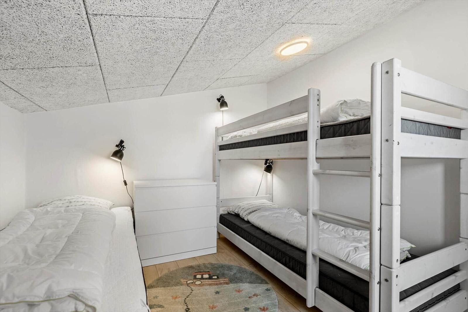 Zimmer mit Doppelstockbett und Einzelbett. Weiße Wände, Holzboden und Deckenleuchte.
